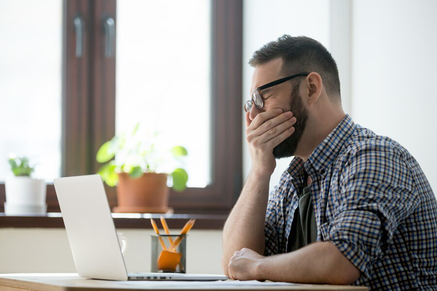 A man in a checkered shirt and glasses sits at his desk in front of a laptop, yawning with his mouth covered by his hand, showing it may be time to shed the extra pounds