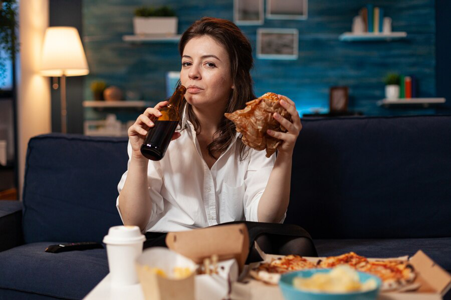 A woman holds a brown glass bottle in one hand and a burger in brown paper in another with pizza, fries, and other junk food laid out in front of her, showing what changes in appetite may look like