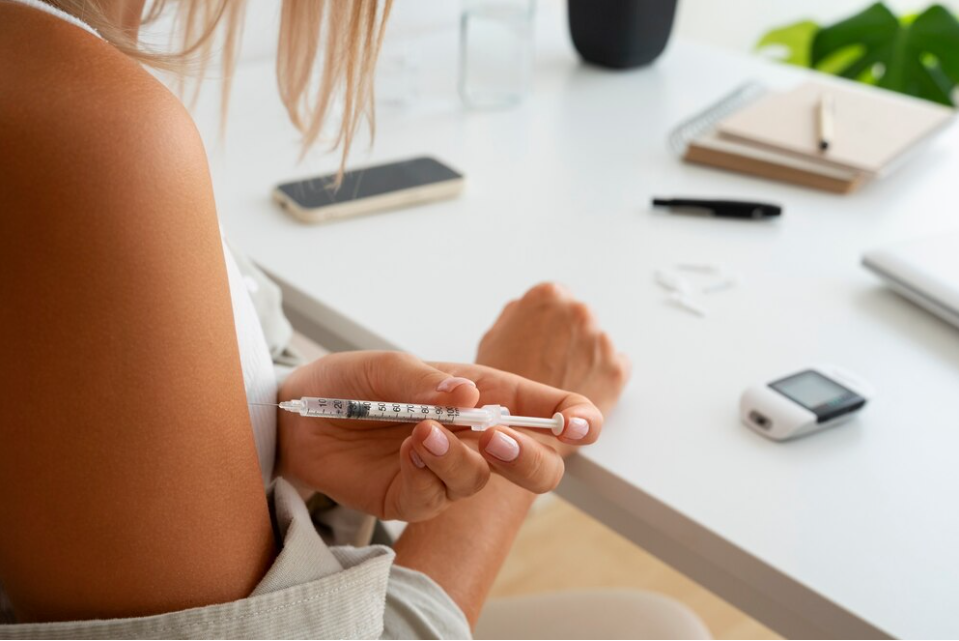 A person injects themselves with an insulin needle in the arm by a desk with a sugar monitoring device and phone