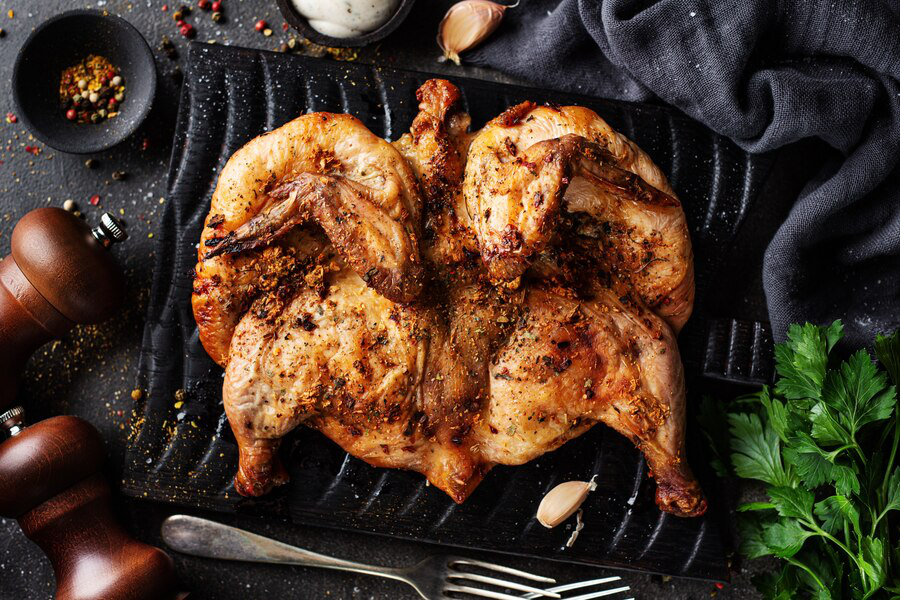 A full butterflied grilled chicken on a black dish alongside cilantro leaves, crushed peppers, forks, and a salt and pepper mill, showing how high protein foods can help with changes in appetite