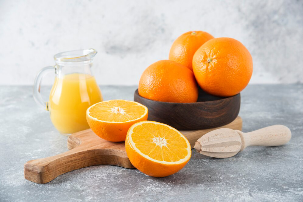 Three oranges in a wooden bowl atop a chopping board with a sliced orange and wooden juicing device along with a small jug of orange juice showcasing how Vitamin C injections can be a quicker, more efficient way of getting nutrients into the body.