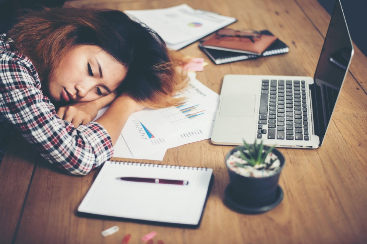 A woman rests her head on her folded arms on her desk amidst paperwork, a laptop, an open notebook, a pen, and a small plant with her eyes shut