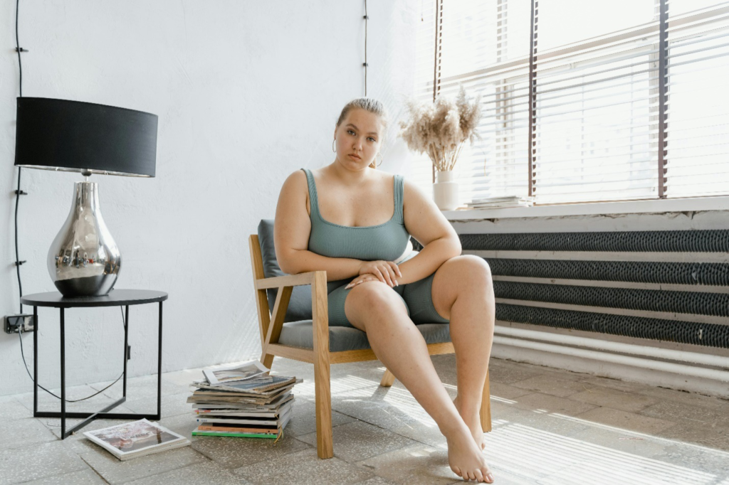 A woman in a grey bodysuit sits on a chair beside a pile of magazines and a black table with a silver and black lamp in front of a window with sunlight streaming in