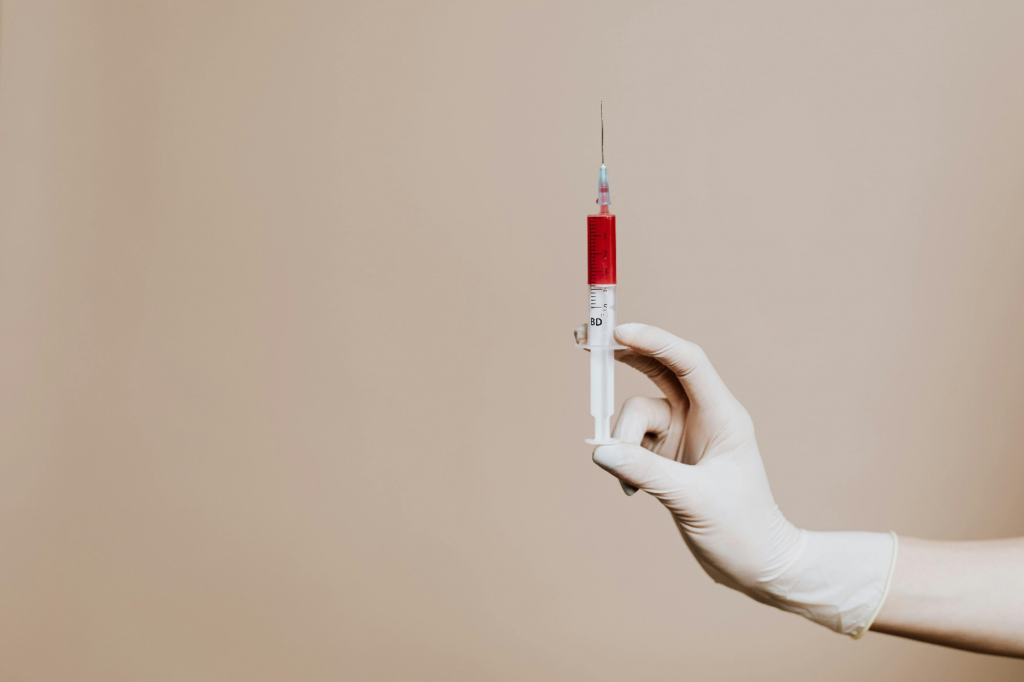 A gloved hand holds a syringe with a needle with a red liquid inside against a grey background