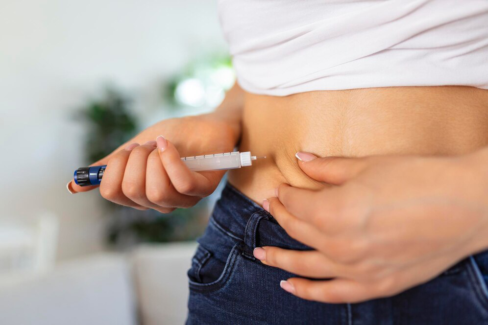 A person in dark indigo jeans and a white t-shirt injects herself in the abdomen with a pen-like injection