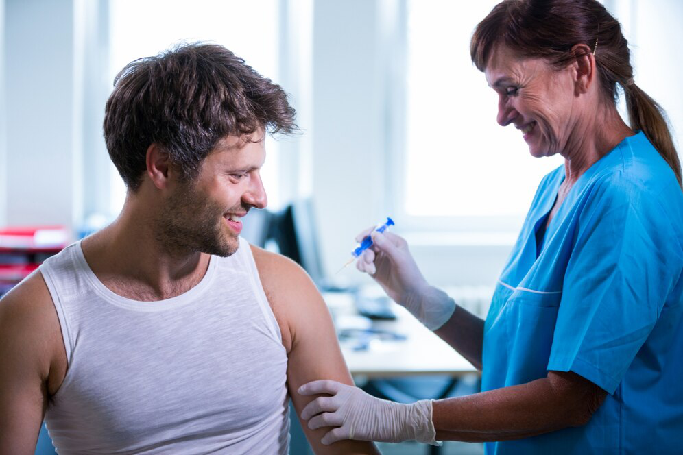 A man in a white vest looks at his arm as a woman in light blue scrubs wearing latex gloves holds an injection in one hand and his arm with the other