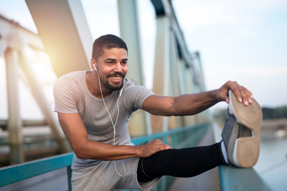 A man stretches his leg while wearing earphones with a bridge in the background