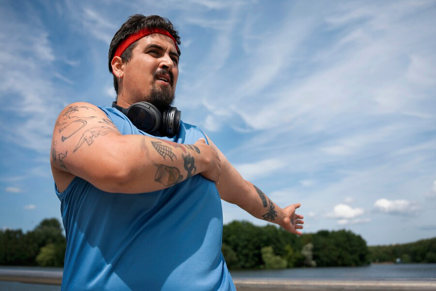 A man in a blue vest and tattoos on his arms wears headphones around his neck outdoors while working out