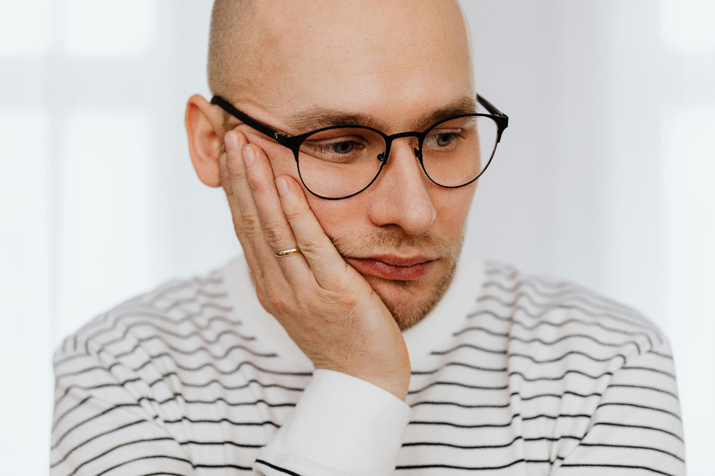 An unhappy looking man in black-rimmed glasses has his hand on his face with a ring on his finger while wearing a black and white full-sleeved t-shirt