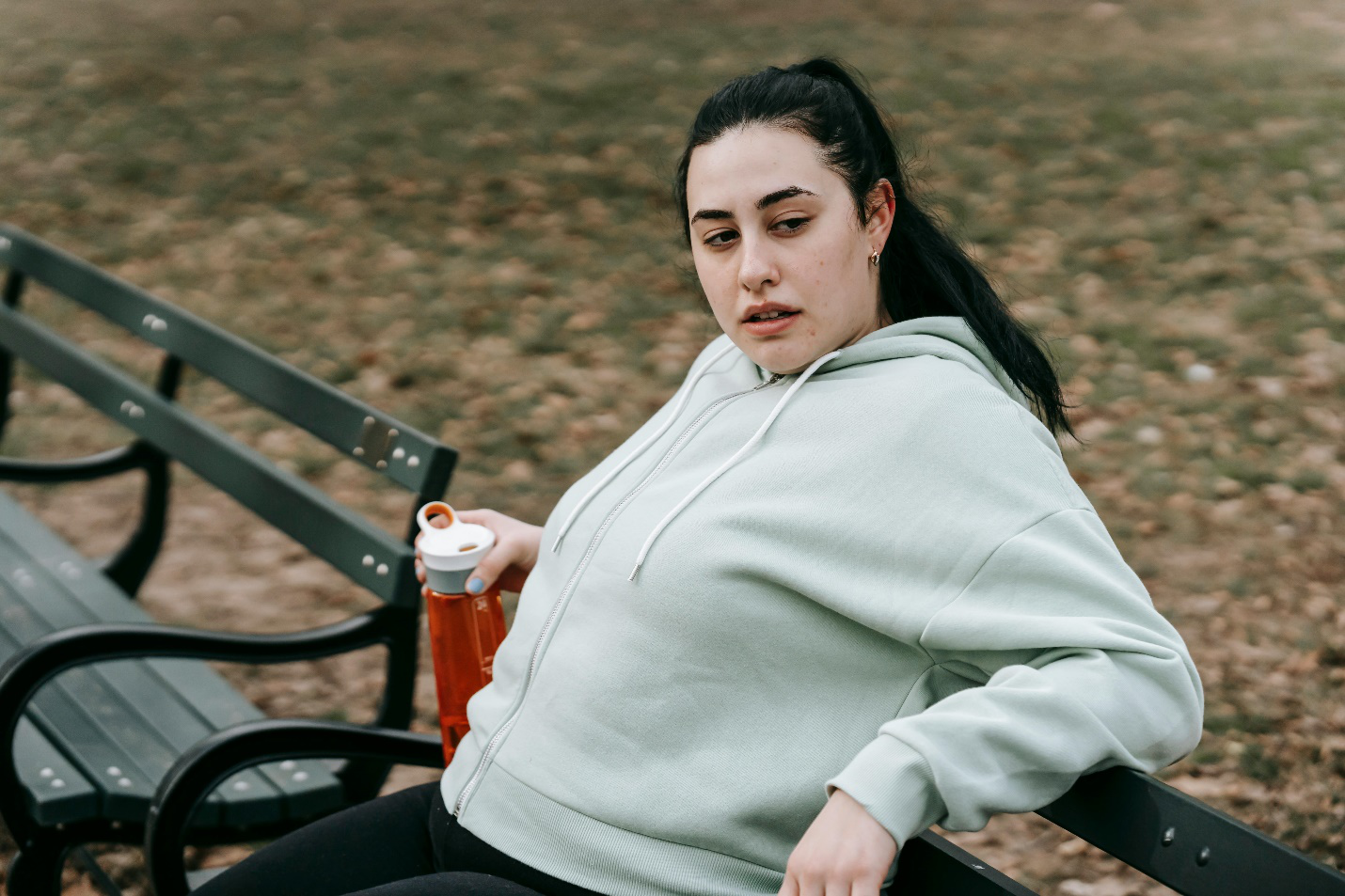 A woman with hair tied back in a light grey jacket and black bottoms sits on a black bench outdoors while holding an orange and white bottle