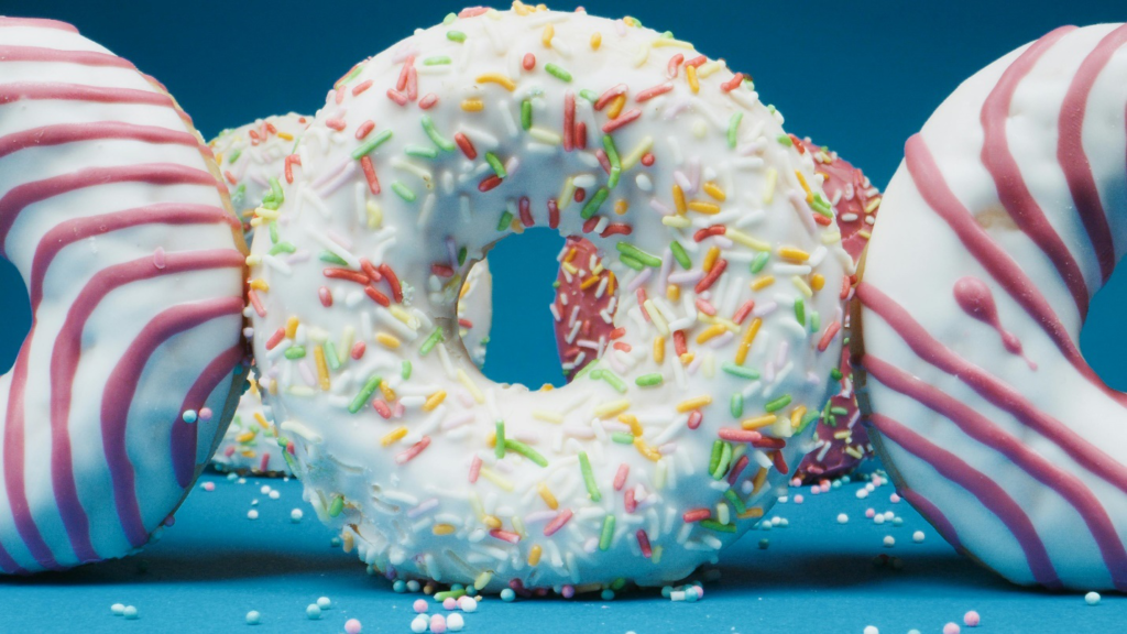 A white sprinkle donut in the middle of two pink and white striped donuts on a blue surface with two sprinkle donuts in the background