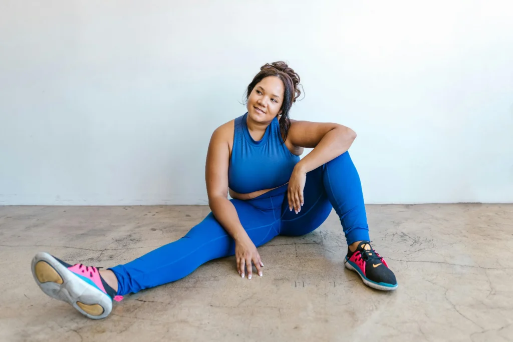 A woman in blue athletic wear and colorful sneakers sits on the ground with one leg extended and the other bent