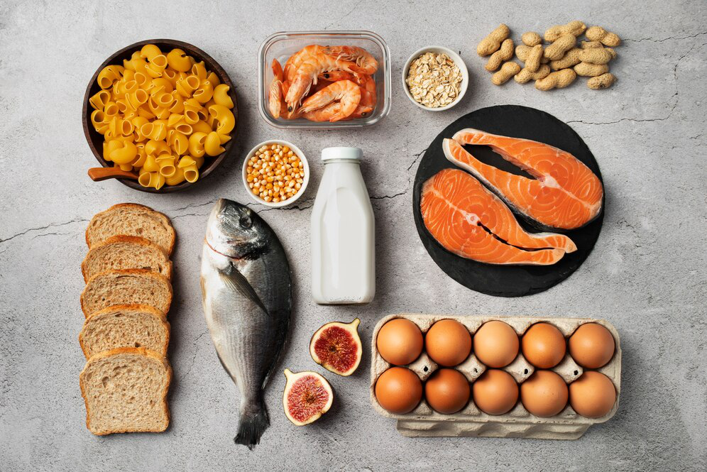 Fish, prawns, eggs, sliced figs, bread, a bowl of raw pasta, peanuts, corn kernels, and oats lie arranged on a textured grey surface showcasing options for ingredients to put together high-protein meals for weight loss