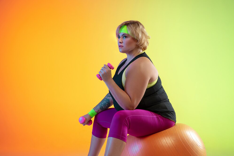  A woman in a black top and magenta tights sits on an orange pilates ball while holding pink dumbells against a bright orange, yellow and lime background