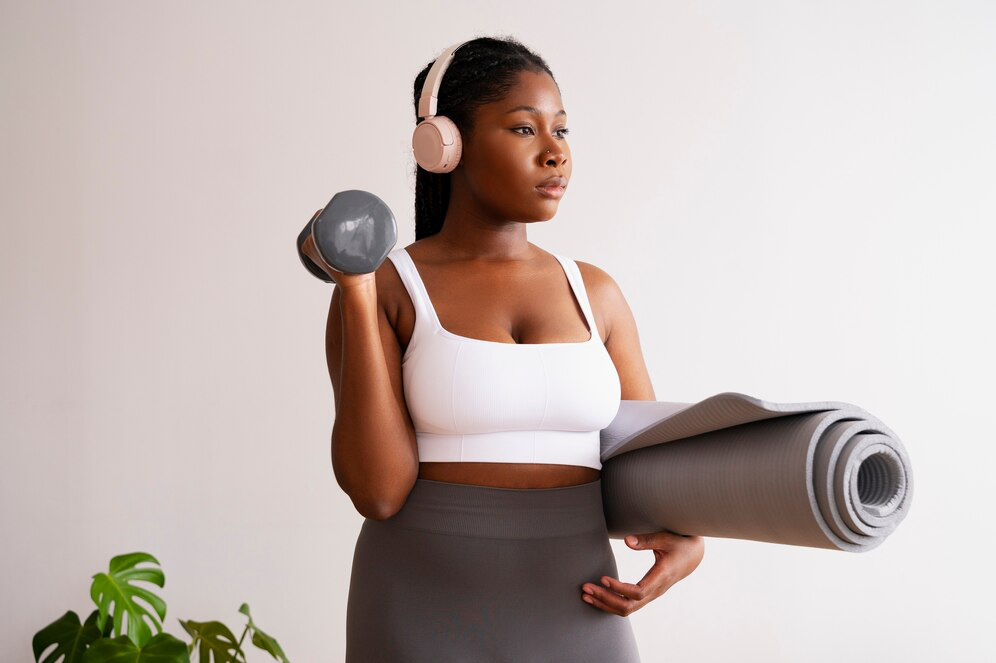 A woman in a white top and grey tights holds a grey yoga mat rolled up and a dumbbell while wearing pink headphones