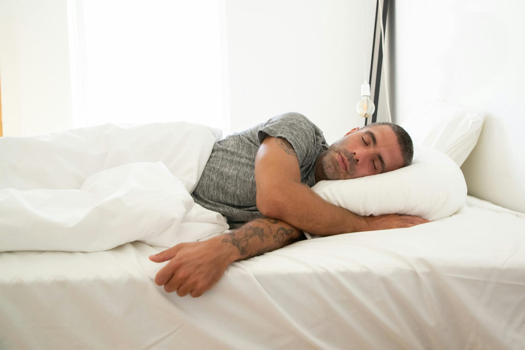 A man with tattooed arms in a grey t-shirt lies in bed under a comforter with his head on a pillow as sunlight streams in