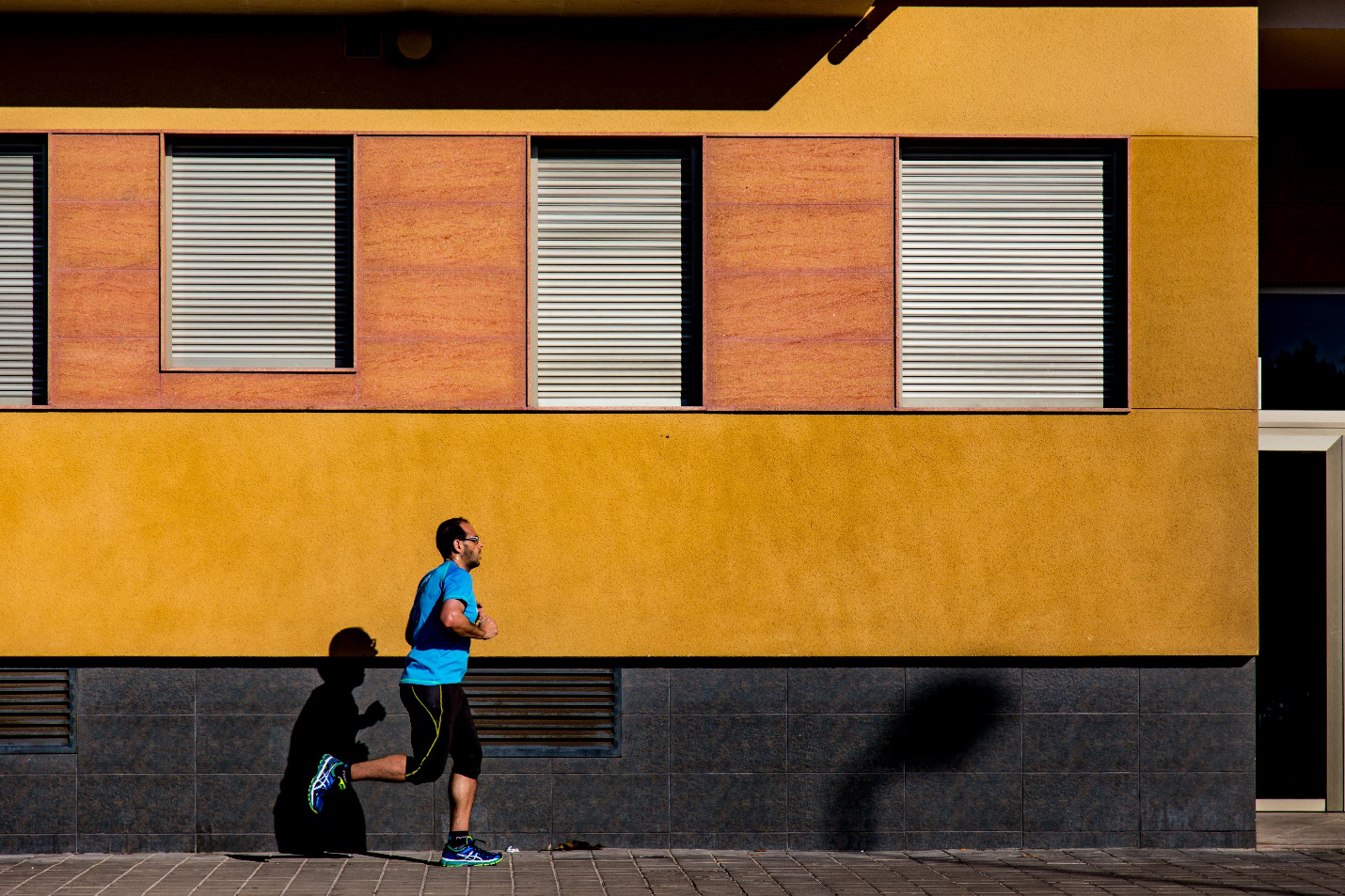  A man in a light blue t-shirt and black shorts runs on the pavement beside a charcoal, ochre, and orange building