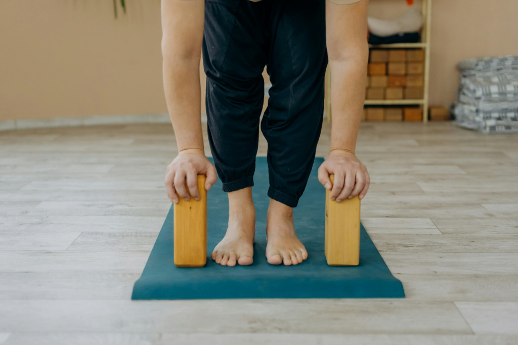 The lower half of a person bent over and resting their hands on two wooden blocks while standing on a blue yoga mat