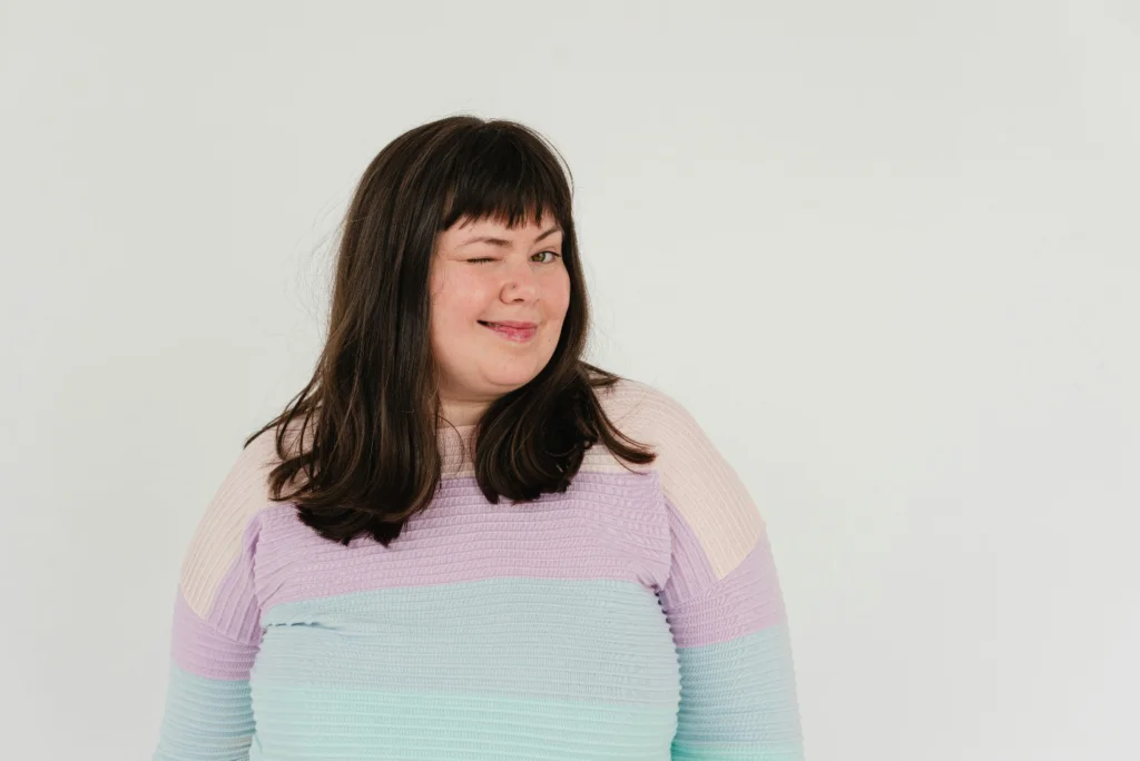 A slightly overweight woman with mid-length hair and a fringe in a pastel color-blocked top winks against a plain white background