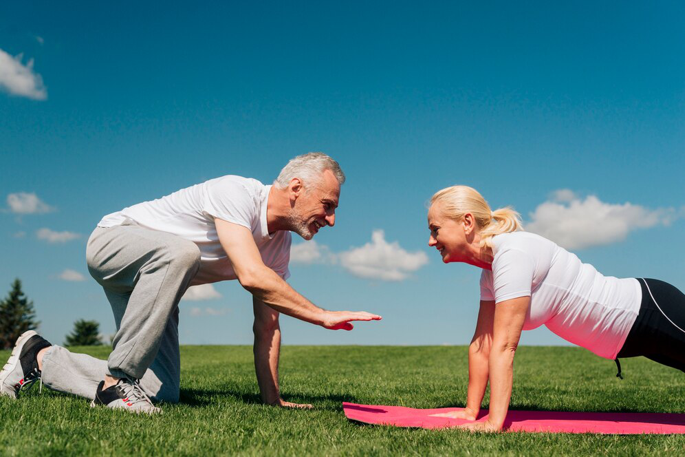 A senior woman in a white t-shirt planks on a pink yoga mat outdoors on the grass while another senior man stands on his knees and hand in front of her