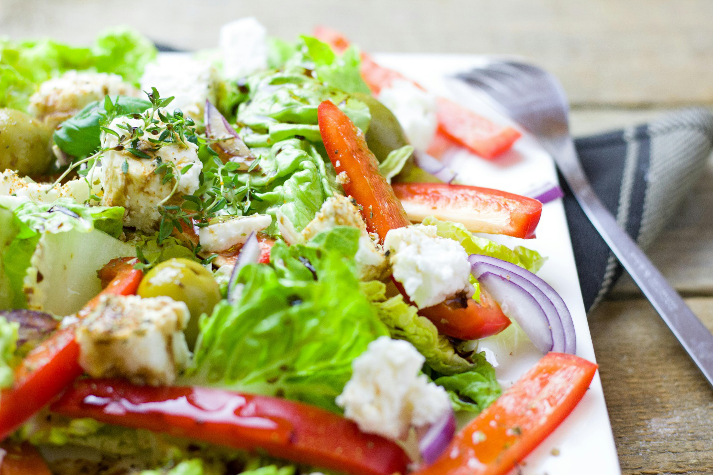 A salad consisting of sliced red peppers, chopped onions, white cheese, hers, olives, and lettuce on a plate with a fork and napkin visible