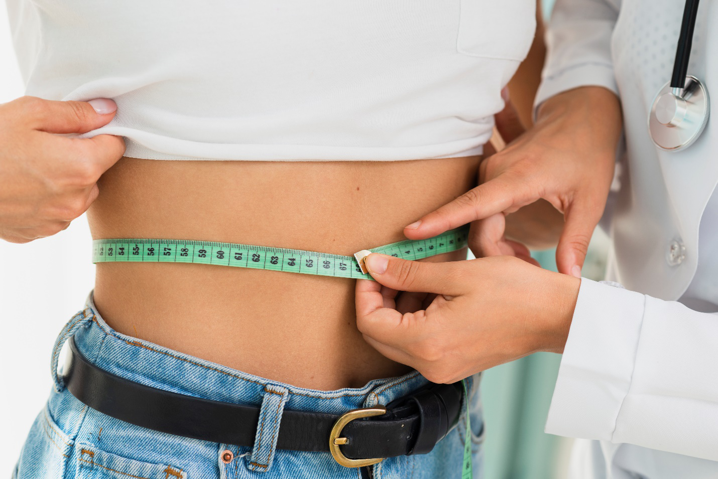 A person wearing a stethoscope uses a light green measuring tape to measure a person’s waist wearing a white t-shirt and blue jeans