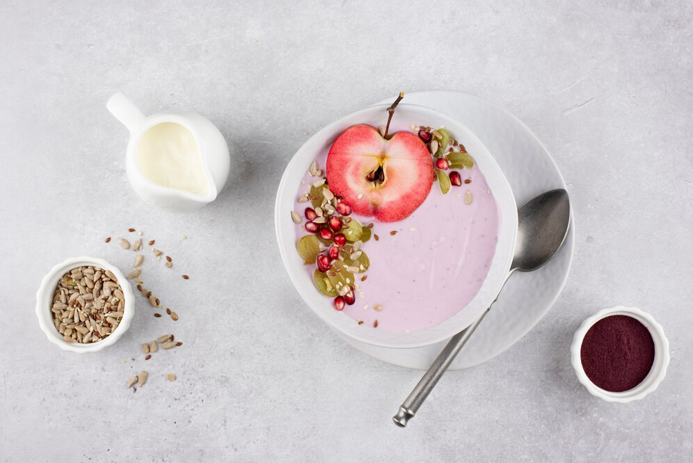 A bowl with a yogurt parfait decorated with fruits and seeds on a saucer with a teaspoon and a small bowl of seeds and milk on a light grey textured surface showcasing an option for a high-protein meal for weight loss