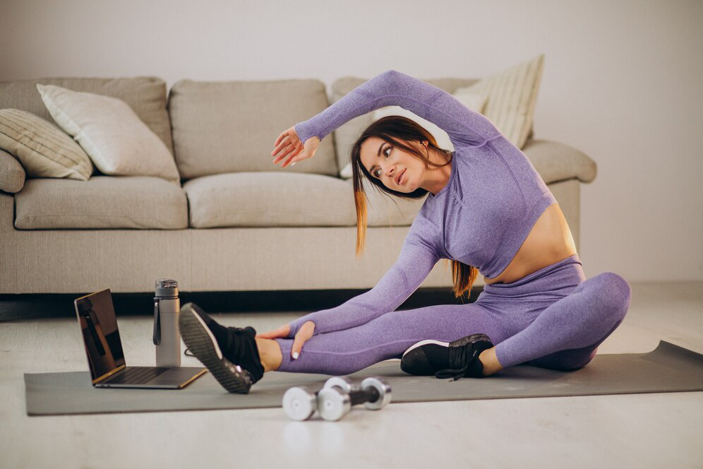 A woman in lavender athletic wear sits on a mat looking at an open laptop with a sofa behind her, bending sideways