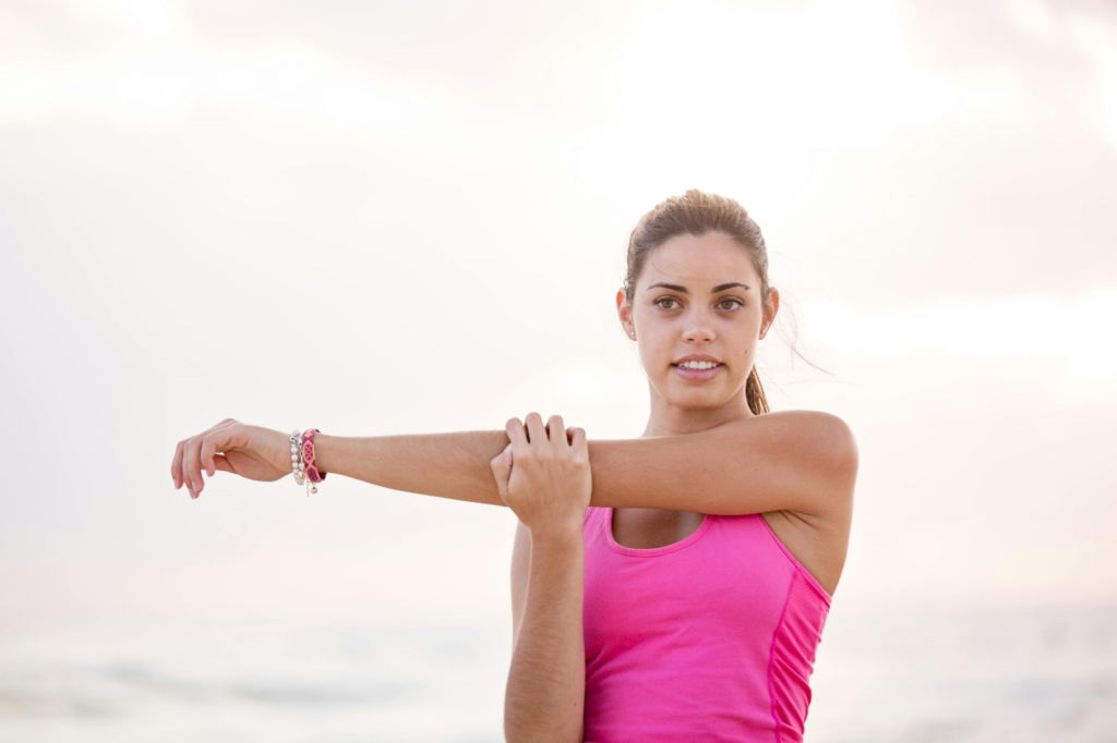 A woman in a pink tank top stretches her arm sideways outdoors with a light pink sky in the background