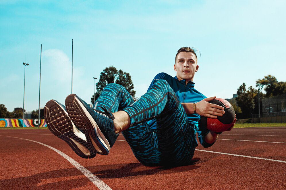 A person in blue athletic wear holds a ball with his legs off the ground, resting on his buttocks on a dark red track surface outdoors