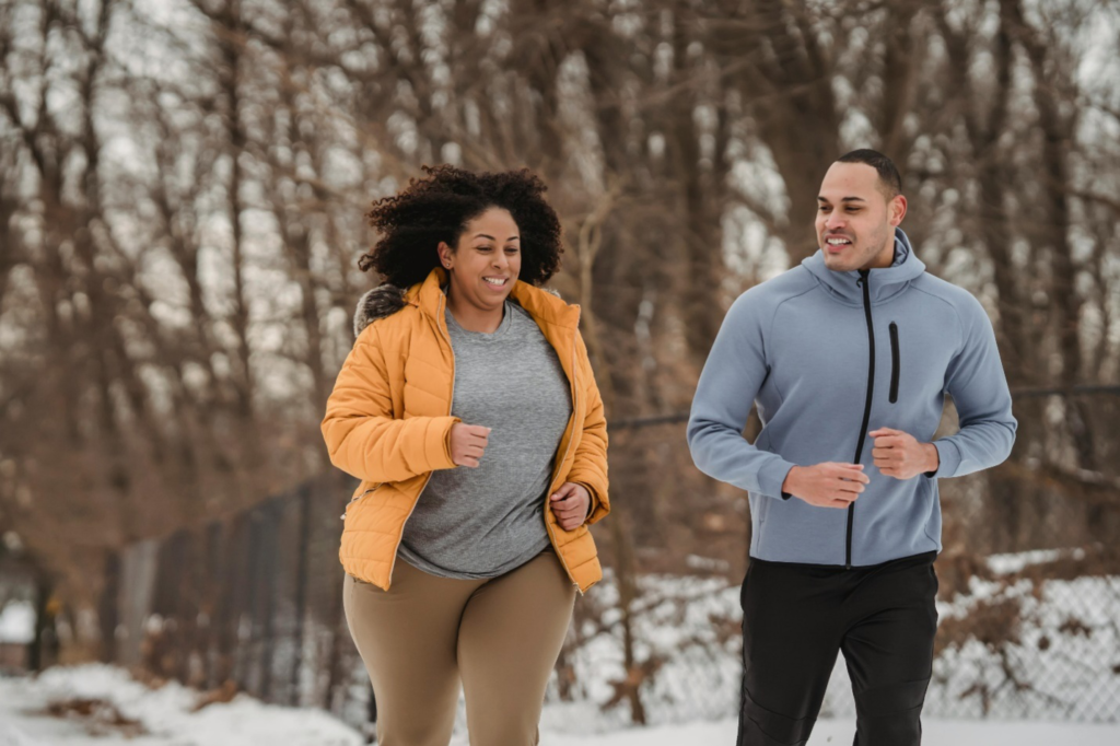 A couple in athletic gear jogs in the snow with barren trees behind them