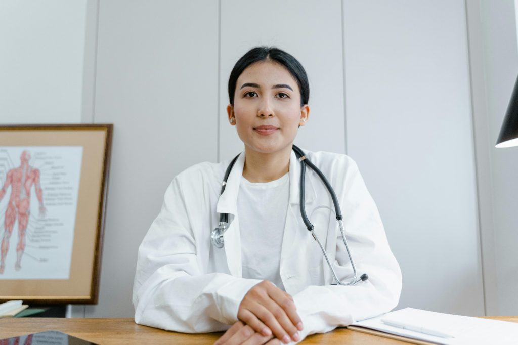 A woman in a white lab coat wearing a stethoscope sits behind a wooden desk with an image of the human body behind her