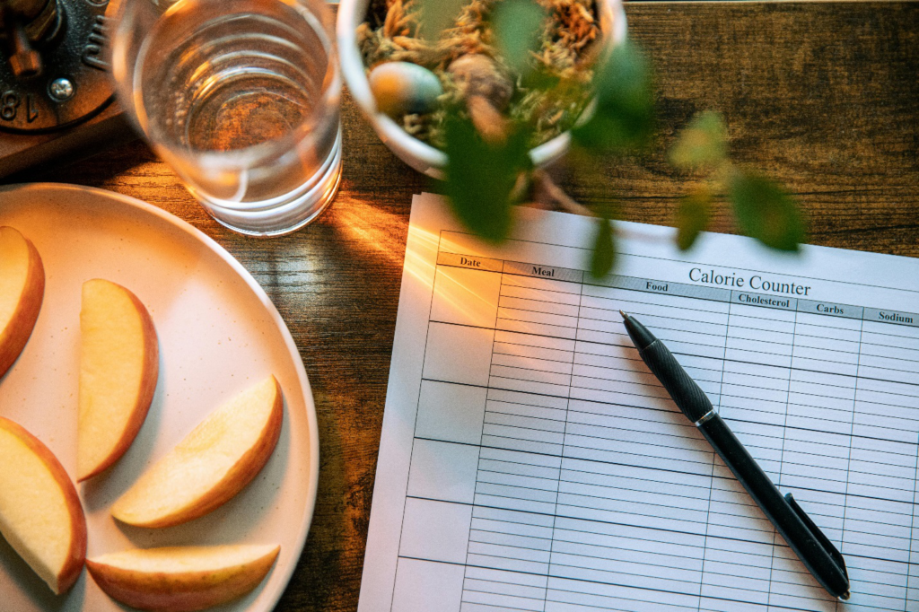 A plate of sliced apples lies by a printed paper reading Calorie Counter with columns for date, meal, food, cholesterol, carbs, sodium, and a pen on top next to a glass of water and planter