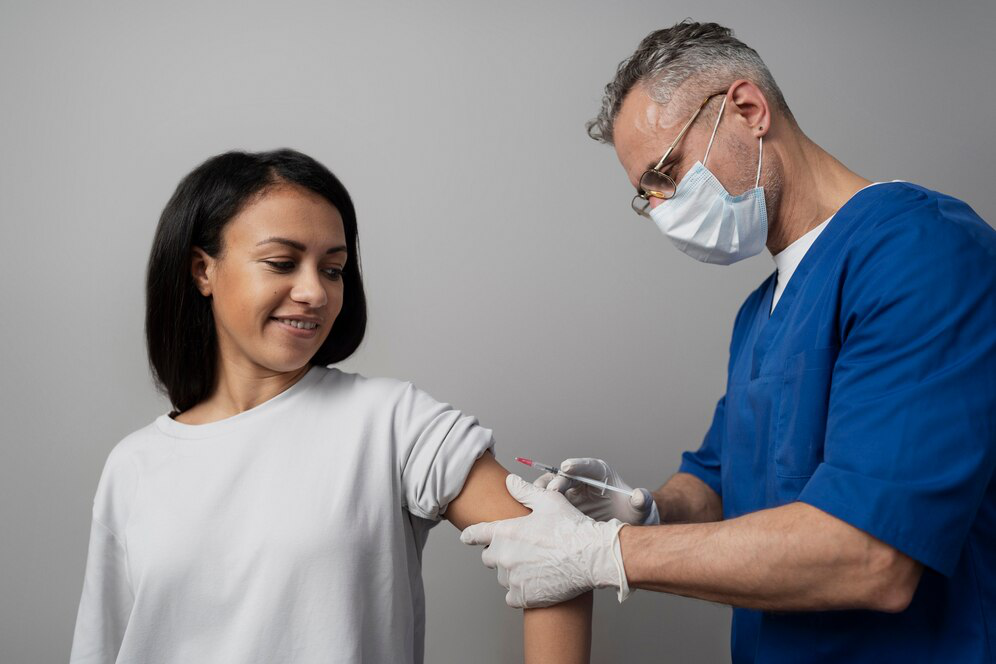  A person in blue scrubs and a mask holds a woman’s arm to administer an injection against a light grey background