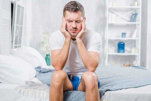 A man sitting on a bed with his head in his hands, looking tired and frustrated.