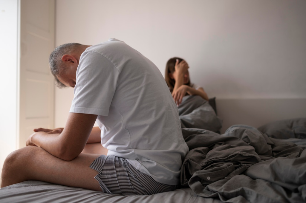 A man sitting on the edge of a bed looking distressed, with a woman sitting behind him in frustration.