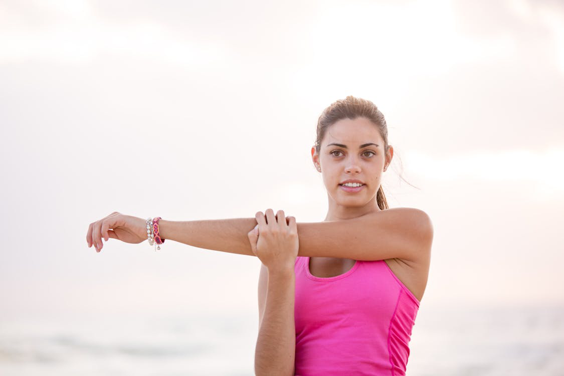 A young woman in a pink tank top stretching on the beach, preparing for a workout.