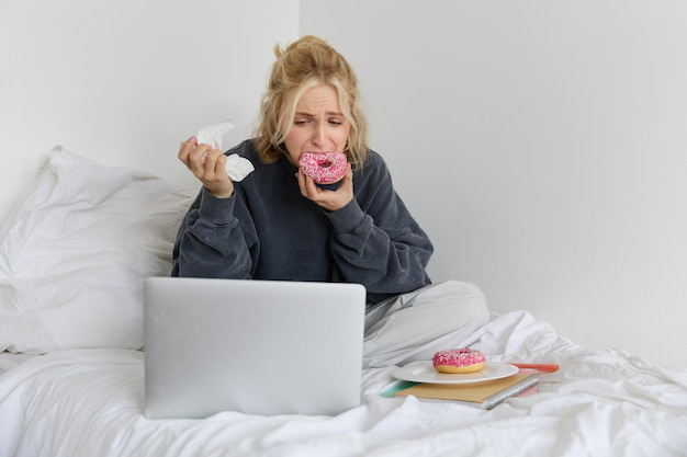 A Woman eating a donut while working on a laptop in bed.