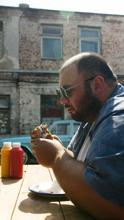 Man sitting outdoors, eating a large sandwich under the sunlight.