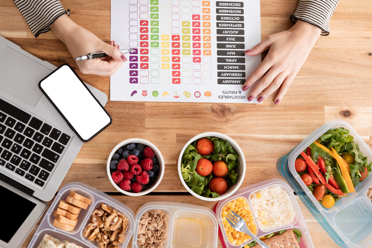 Person tracking nutrition and seasonal produce besides prepped healthy meals and fruits