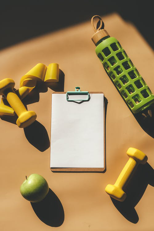 A fitness plan clipboard next to dumbbells, a water bottle, and an apple