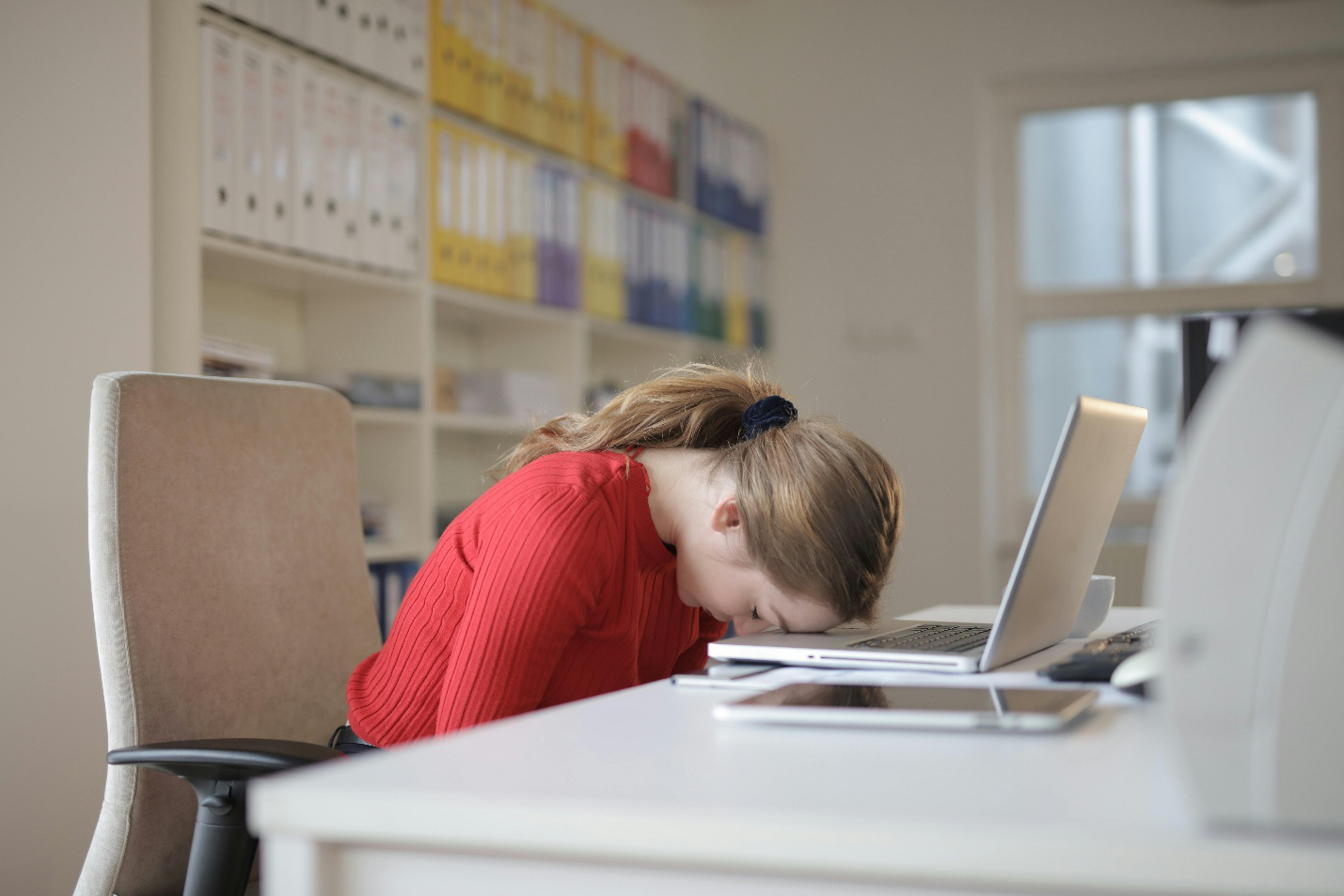Woman in red shirt with head down on desk, showing signs of work-related fatigue