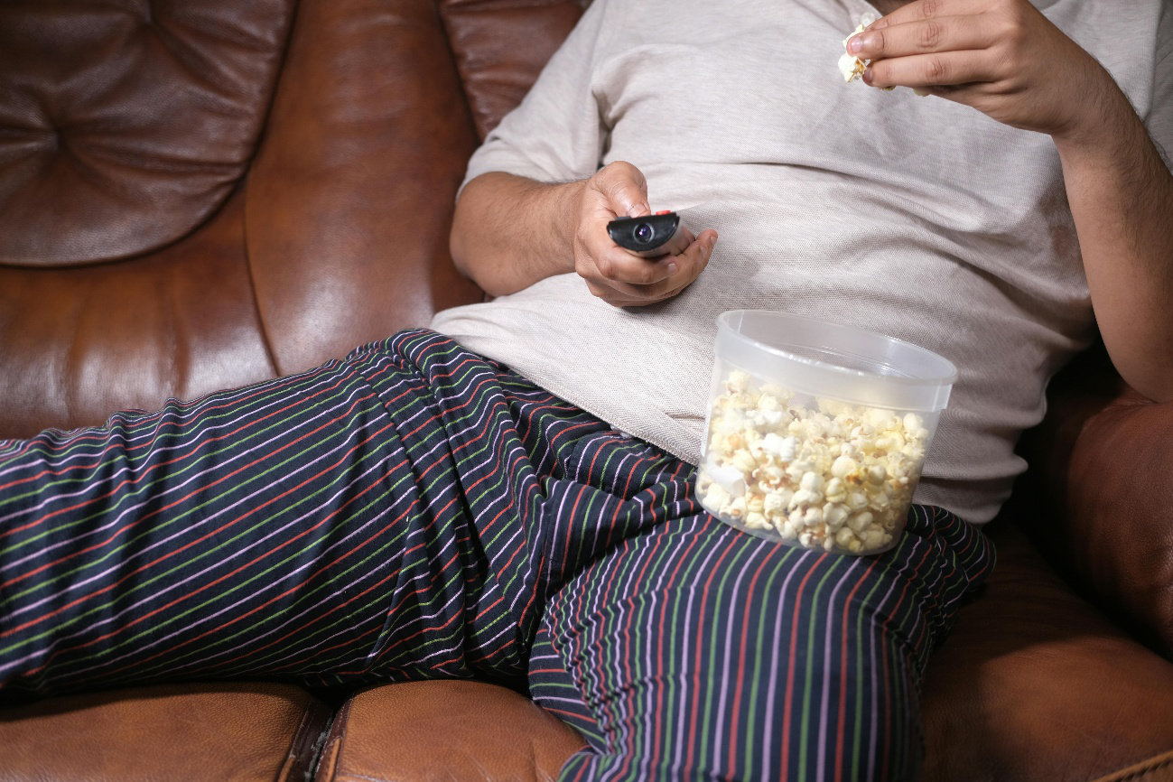Man lounging on a leather couch in pajamas, eating popcorn, and watching TV