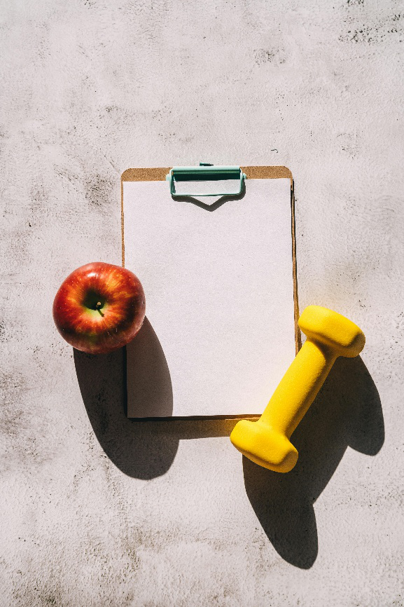 A blank health checklist clipboard with a red apple and yellow dumbbell on a concrete background