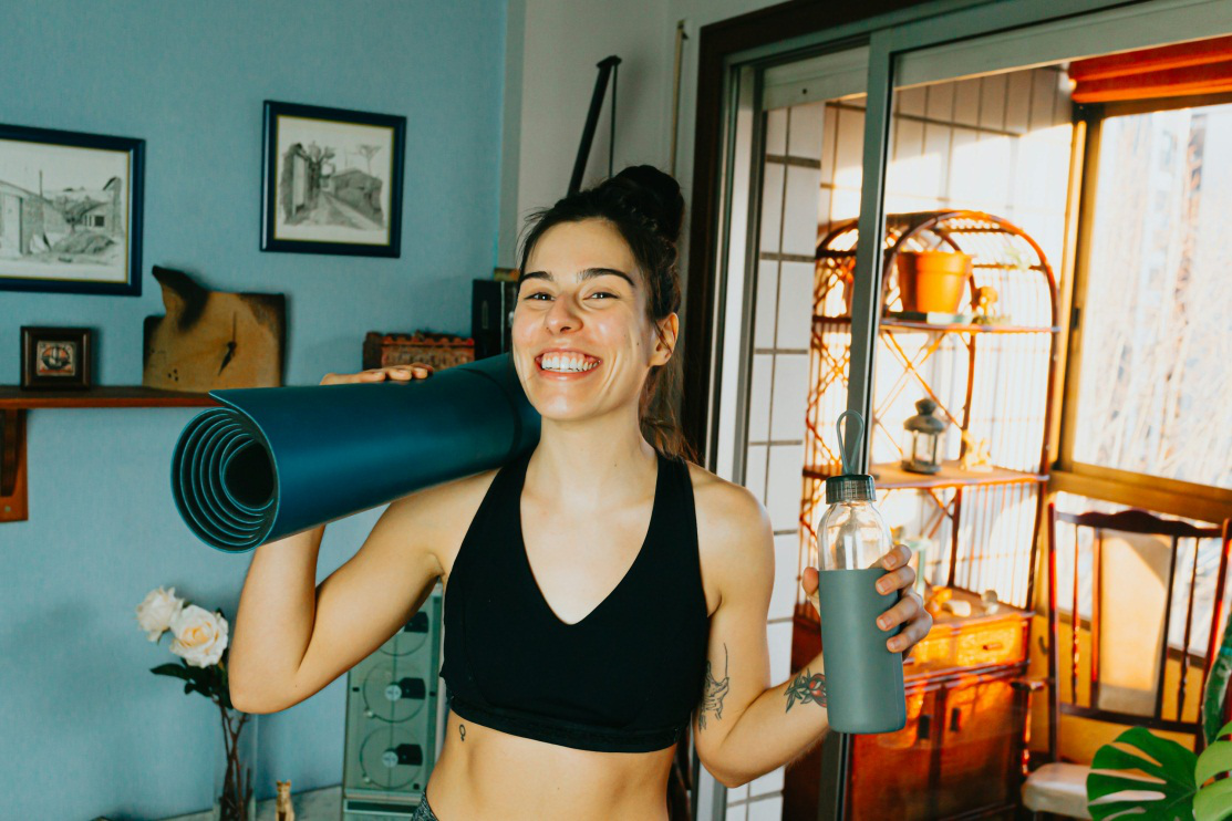 A photo of a smiling woman holding a yoga mat and a water bottle