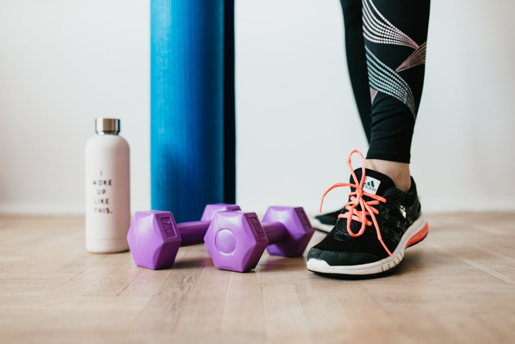 An image of a person standing next to dumbbells, a yoga mat, and a bottle   