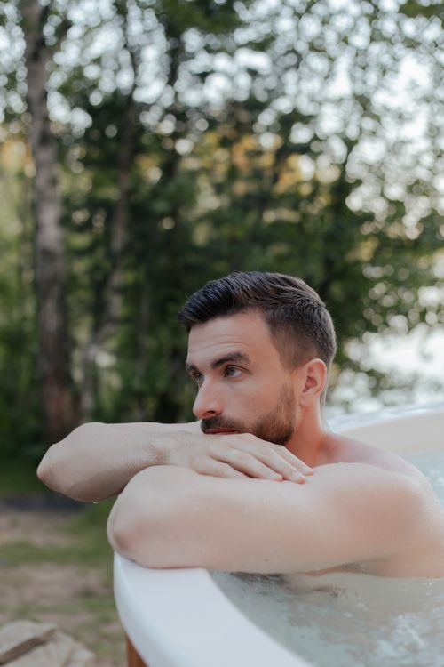  Man resting his arms on the edge of an outdoor hot tub surrounded by trees