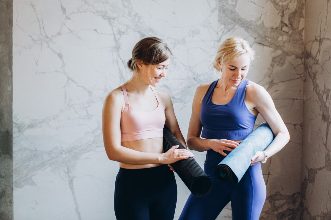 Two women in workout clothes holding rolled-up yoga mats while talking