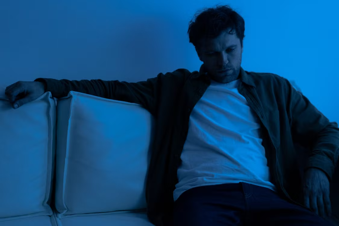 Man sitting on a couch in a dimly lit blue-toned room looking down