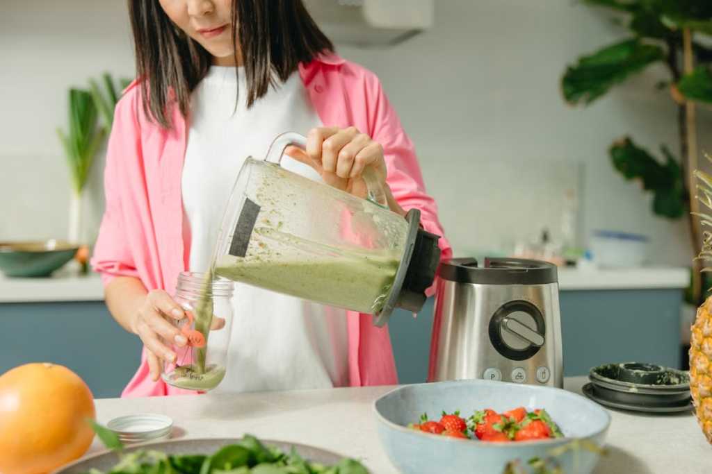 Woman in a pink shirt pouring a green smoothie from a blender into a glass jar in a kitchen
